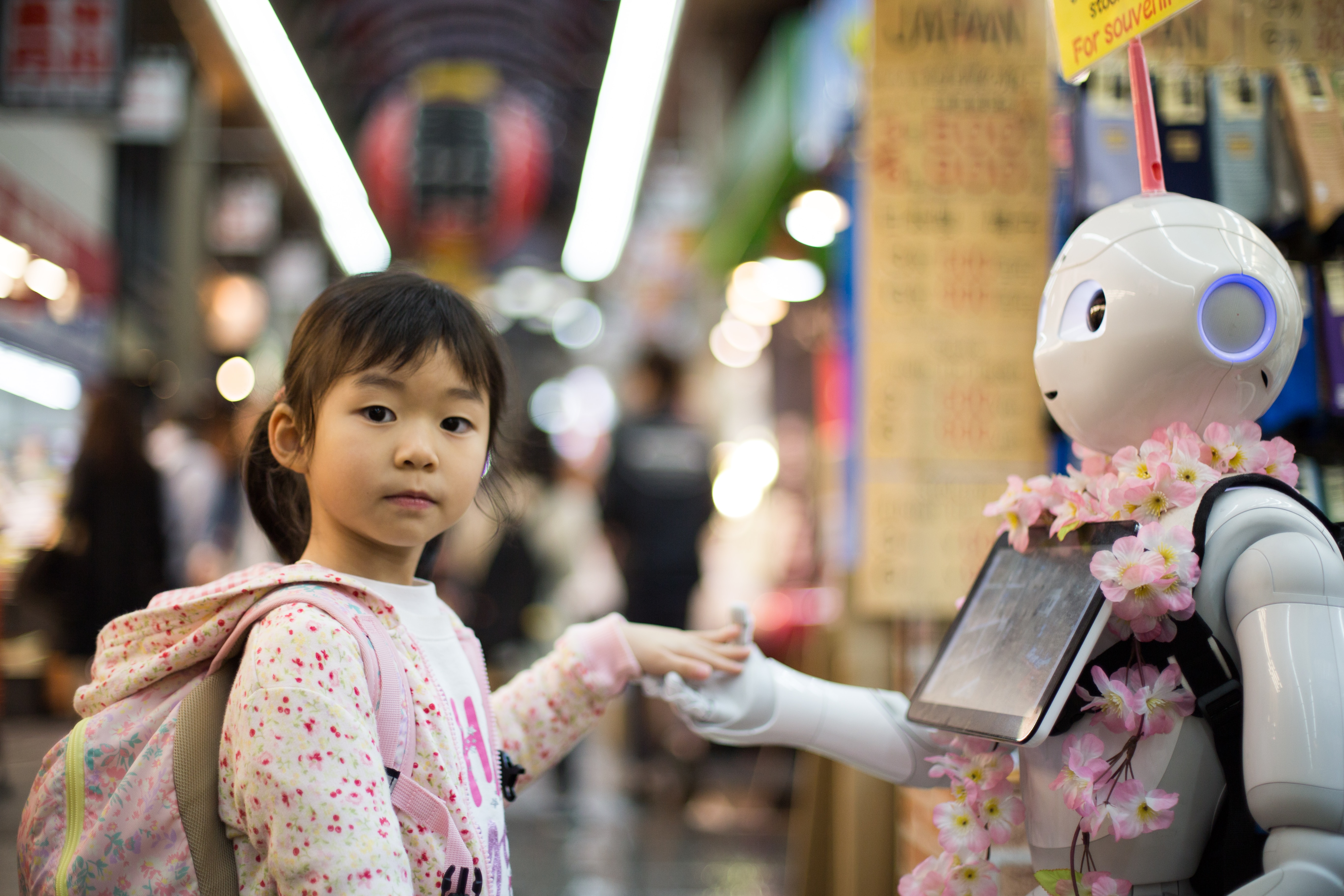 little-girl-holding-the-hand-of-a-similarly-sized-robot-wearing-a-pink-flower